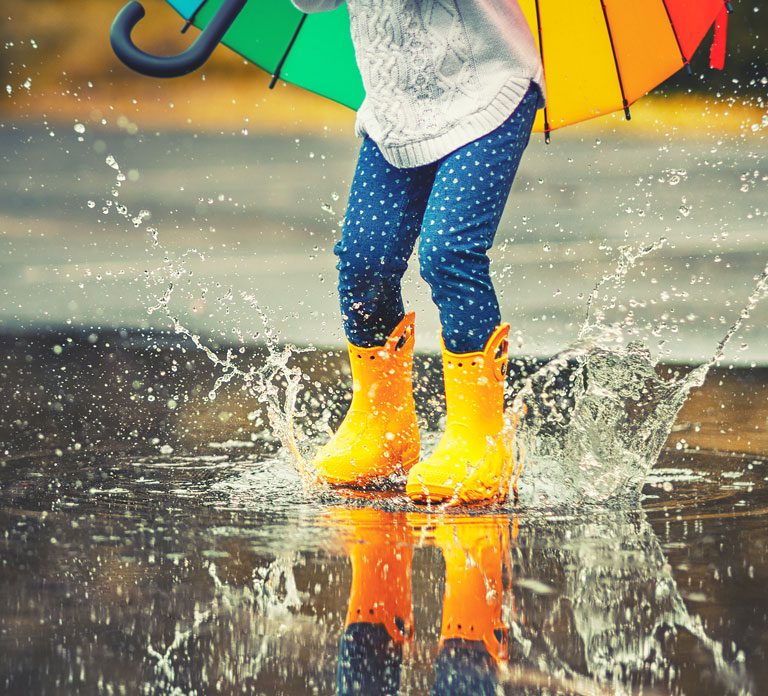 child with rainbow umbrella wearing yellow rain boots playing in puddle